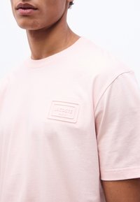 Νεαρό άτ ομο που φοράει ροζ ανοιχτό μπλουζάκι με ένα ανυψωμένο ορθογώνιο έμπλαστρο που γράφει "Lacoste Paris" στο στήθος.