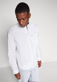 Camicia bianca a maniche lunghe in cotone, con taschino sul petto e un piccolo logo. Maniche lunghe con polsini abbottonati, design del colletto classico.