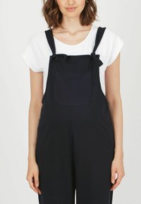 Marinblå hängselbyxor i lätt tyg, med justerbara remmar, en framficka och löst sittande ben. Kombinerat med en vit T-shirt.