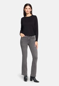 Pull noir à manches longues associé à un jean gris droit. Le mannequin porte des bottines noires à talons et pose avec une main sur la hanche.