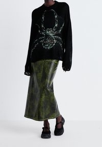 Zwarte gebreide trui met groene spinnenafbeelding, gecombineerd met een lange, glanzende, groen leren rok. Schoenen hebben een zwart textuur ontwerp.