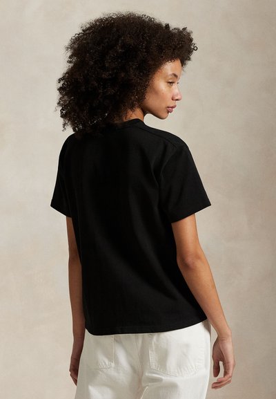 Femme aux cheveux bouclés portant un t-shirt noir à manches courtes et un pantalon blanc, de profil devant un fond beige uni.