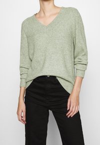 Vaaleanvihreä, ribboitunut neulepusero, jossa on V-pääntie ja käännetyt hihansuut, yhdistettynä mustiin housuihin. Pehmeä rakenne, rento istuvuus, sivuhihoissa halkiot.