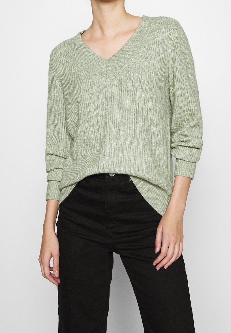 Vaaleanvihreä, ribboitunut neulepusero, jossa on V-pääntie ja käännetyt hihansuut, yhdistettynä mustiin housuihin. Pehmeä rakenne, rento istuvuus, sivuhihoissa halkiot.