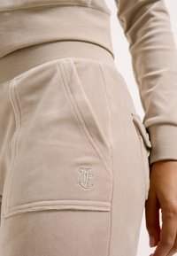 Beige velours sweatpants met een aangesloten tailleband, zakken en geborduurd logo op de zijkant. Soepele textuur, op maat gemaakte stiksels.