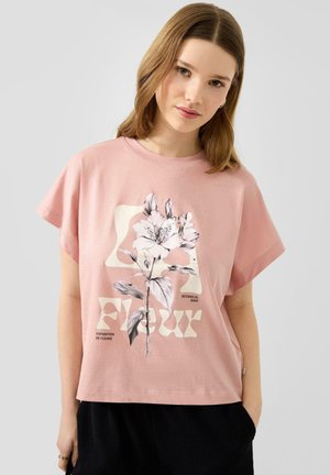 Jonge vrouw met lichtbruin haar draagt een loszittend roze T-shirt met een groot bloemmotief en tekst, gecombineerd met een zwarte broek.
