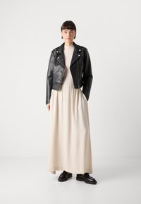 Black leather biker jacket over a light beige maxi dress with a textured finish, paired with black loafers. Plain background.