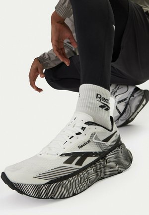 Persona che indossa scarpe da ginnastica Reebok bianche e nere con suole decorate e calzini coordinati con il marchio, accovacciata su uno sfondo bianco.