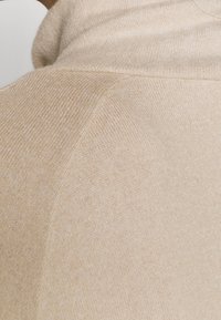 Pull en maille beige à texture lisse, avec un col montant et une coupe ajustée. Des détails de coutures sont visibles le long de l'épaule.