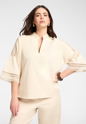 Kobieta o ciemnych włosach w kremowej bluzce z koronkowymi detalami na rękawach i dopasowanych spodniach, uzupełniona złotymi kolczykami i bransoletką.