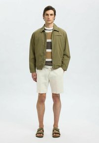 Olivově zelená bomber bunda, pletený svetr s pruhy v hnědé, krémové a béžové, béžové šortky a tmavé sandály. Jednoduchý design, ležérní vzhled.