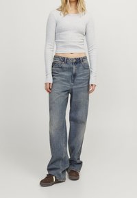 Eine Frau trägt ein hellgraues langärmliges Cropped-Top, lockere ausgebleichte blaue Jeans und braune Sneaker und steht vor einem schlichten weißen Hintergrund.