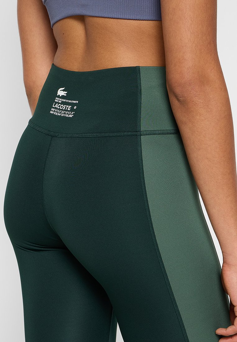 Leggings de sport taille haute de couleur vert foncé, fabriqués en matériau extensible. Présente un panneau latéral contrastant et un logo de marque à la taille.