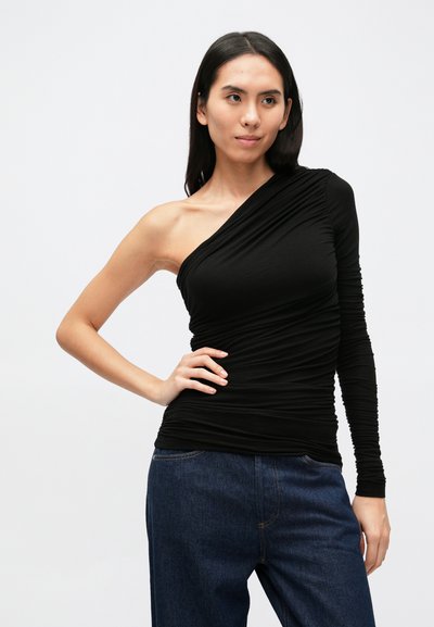 Top preto de manga comprida com um ombro exposto, apresentando um design franzido para textura. Combinado com jeans azul escuro.