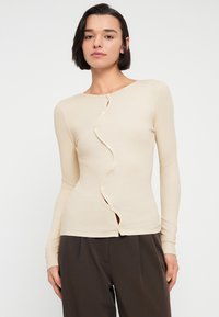 Μπεζ μπλούζα με μακριά μανίκια, με κυματιστό μπροστινό μέρος με κούμπωμα και ριμπ υφή. Συνδυασμένη με σκούρο παντελόνι, τονίζοντας το εφαρμοστό σχέδιο.