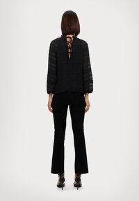 Top in pizzo nero con maniche a sbuffo, schiena scoperta con fiocchi, indossato con pantaloni neri a zampa d'elefante e scarpe nere con tacco.