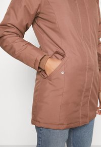 Brązowa, ocieplana kurtka o gładkiej fakturze, z zamkiem błyskawicznym z przodu, bocznymi kieszeniami i ściągaczami. Stylizowana z jasnoniebieskimi dżinsami.