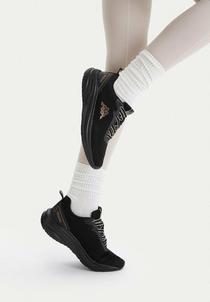Czarne sportowe buty z siateczki z białymi sznurowadłami, noszone z białymi prążkowanymi skarpetkami i beżowymi legginsami na jednolitym białym tle.