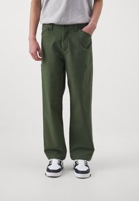 Green cotton pants with a straight fit, featuring two front pockets, belt loops, and a button closure. Worn with black and white sneakers.