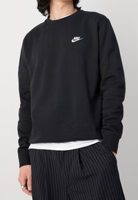 Černá mikina Nike s kulatým výstřihem, dlouhými rukávy a bílým logem na hrudi. Nositá přes bílou košili, v kombinaci s kalhotami s proužky.