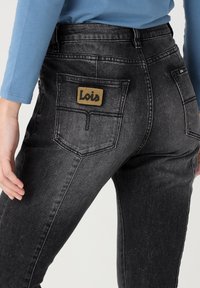 Dark gray denim jeans featuring a high waist, two back pockets, and a tan logo patch on the waistband. Smooth texture with slight stretch.