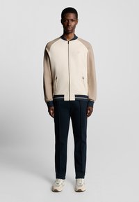 Herre bomberjakke i creme og beige med lynlås, ribbede manchetter og sidelommer. Sammen med navy tapered bukser og lyse sneakers.