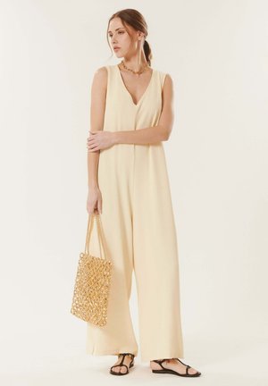 Femme portant une combinaison crème sans manches et des sandales noires, tenant un sac à main tressé doré, debout devant un fond clair uni.