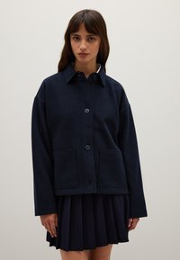 Navy blue wool blend jacket with a collar, front button closure, and patch pockets; worn over a pleated navy skirt. Soft texture.