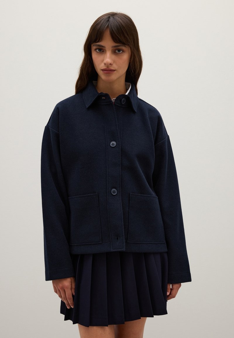 Navy blue wool blend jacket with a collar, front button closure, and patch pockets; worn over a pleated navy skirt. Soft texture.