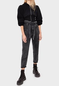 Felpa nera con zip e maniche a sbuffo, abbinata a jeans neri a vita alta, modello largo, con cintura annodata e lunghezza alla caviglia. Indossata con stivali neri robusti.