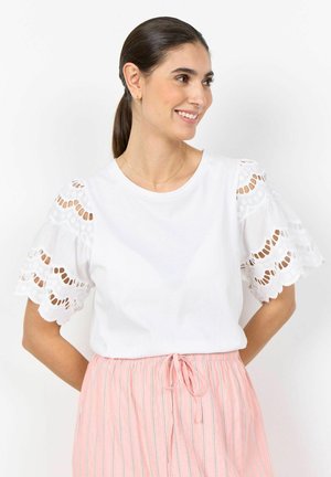 Tumejuustega naine, kes kannab valget lühikeste varrukatega silmkoelist pluusi ja roosade triipudega seelikut, naeratab ja vaatab kõrvale.