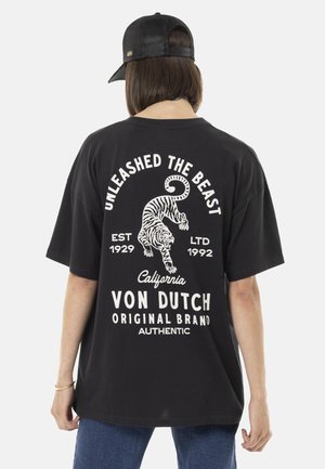 Personne portant une casquette noire et un T-shirt noir ample avec un texte blanc et un graphisme de tigre indiquant « Libérez la bête, Von Dutch, Californie ».