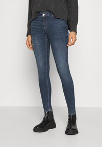 Una persona está de pie vestida con jeans ajustados de color azul oscuro y un suéter gris, combinados con botas de tobillo negras, frente a un fondo blanco liso.