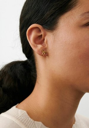 Közeli kép egy személy füléről, akinek sötét haja hátra van fogva, és aki egy kis, absztrakt formájú arany fülbevalót visel.