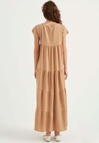 Maxikleid in einem hellbeige Farbton mit Kurzärmeln, charakterisiert durch ein gestuftes Design und einen strukturierten Stoff. Fällt bis zur Knöchellänge.