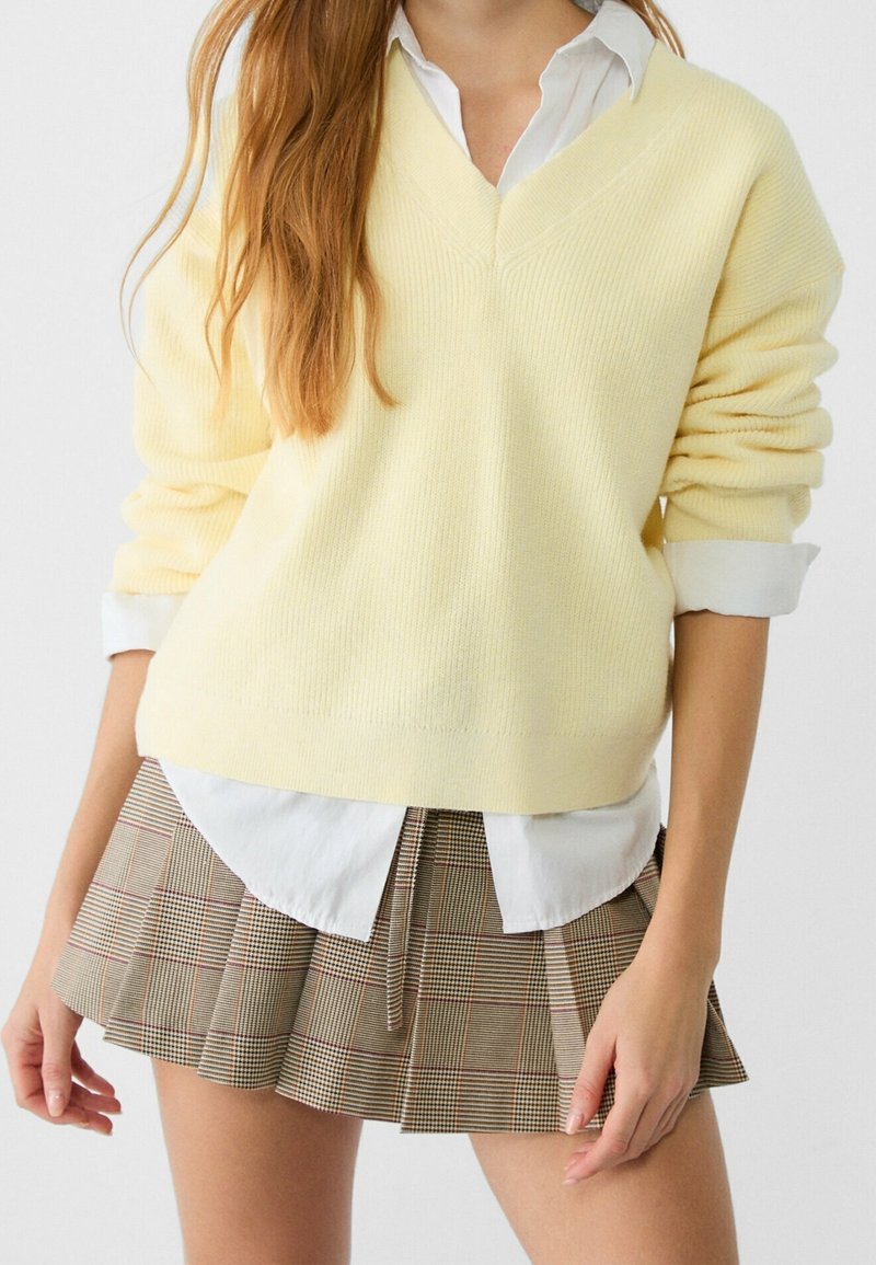 Pull en maille jaune avec un décolleté en V superposé sur une chemise blanche à col, associé à une jupe plissée écossaise marron.