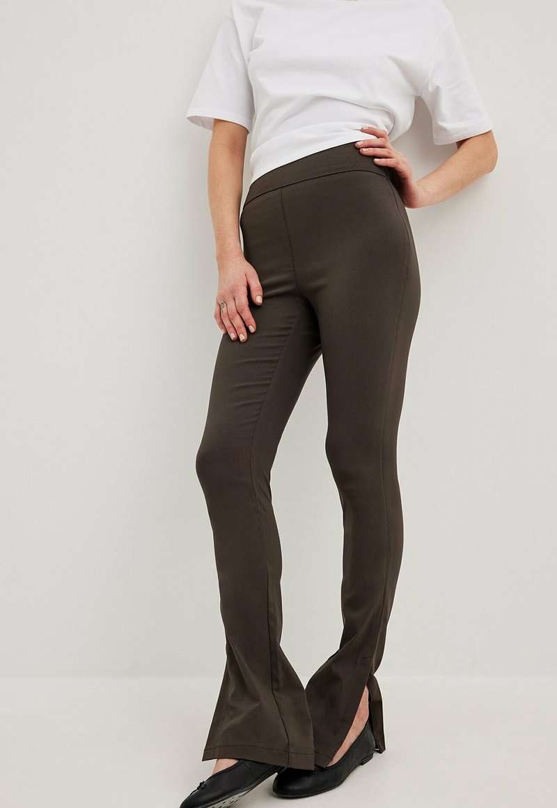 Personne portant un pantalon marron foncé ajusté avec des fentes à l'avant des jambes, associé à des ballerines noires et une chemise blanche à manches courtes, debout contre un mur uni.