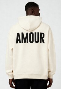 Vyras, atsisukęs nugara, vilkintis kreminės spalvos hoodie su dideliu juodu tekstu "AMOUR" ant nugaros ir juodomis kelnėmis, priešais vientisą foną.