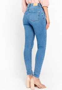 Kvinna som bär ljusblå skinny jeans och blommig rosa klacksandaler, står med ena handen vilande på sin sida.