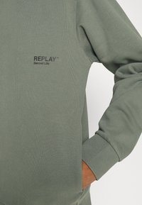 Sudadera verde con textura acanalada, que presenta un cuello redondo, mangas largas y un logotipo bordado que dice "REPLAY Second Life" en el pecho.