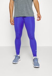 Niebieskie legginsy sportowe wykonane z elastycznego materiału, o gładkiej fakturze i dopasowanym kroju, zestawione z melanżową szarością T-shirtem i szarymi trampkami.