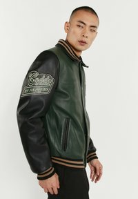 Veste bomber en cuir vert avec manches en cuir noir, col côtelé et poignets rayés. Dotée d'un logo brodé sur la manche gauche.