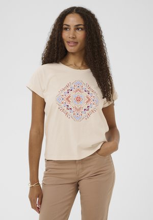 Kobieta o długich kręconych włosach, ubrana w beżowy t-shirt z wielokolorowym geometrycznym wzorem i jasnobrązowe spodnie, ręka w kieszeni.