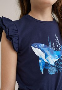 Námořnické modré tričko s grafikou modré velryby a mořských prvků. Volánkové rukávy dodávají texturu a detail designu.