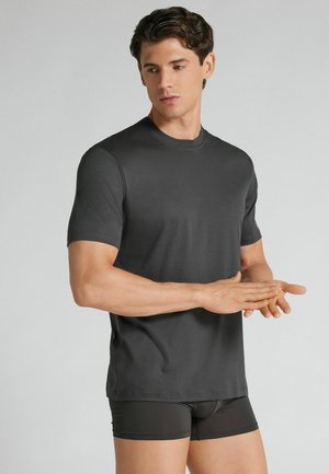 Man draagt een aansluitend donkergrijs T-shirt met korte mouwen en donkere boxershorts, staat met gevouwen handen en kijkt iets opzij.