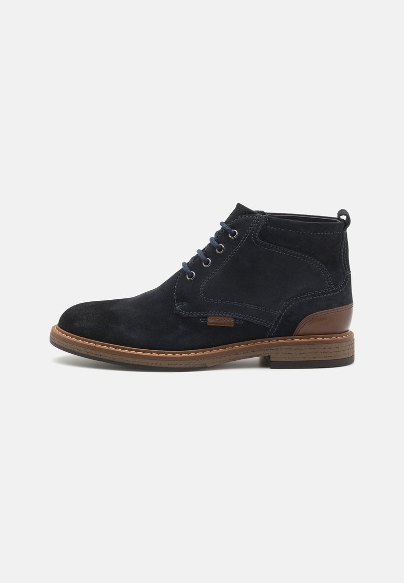 Bottine en daim noire avec détail de talon en cuir marron, lacets foncés et semelle cousue sur un fond clair uni.