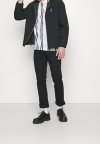 Svart lättviktsjacka med huva, vit randig skjorta, svarta byxor och svarta skor med tjocka sulor. Avslappnad passform med elastiska ärmslut.