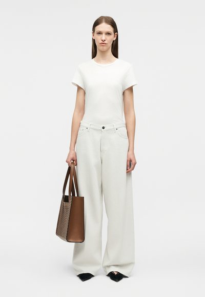 Γυναίκα που φοράει λευκό t-shirt και φαρδιά λευκά παντελόνια, κρατώντας καφέ τσάντα tote, στέκεται μπροστά σε ένα απλό ανοιχτό φόντο.