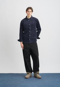 Σκούρο μπλε καρώ πουκάμισο με κουμπιά και γιακά, σε συνδυασμό με φαρδιά μαύρα παντελόνια και μπεζ μποτάκια μέχρι τον αστράγαλο σε μπλε μοκέτα.