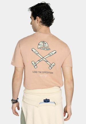 Hombre que lleva una camiseta melocotón con martillos de camuflaje cruzados y un gráfico de gorra de camuflaje y el texto "Lead the Expedition" en la parte trasera.